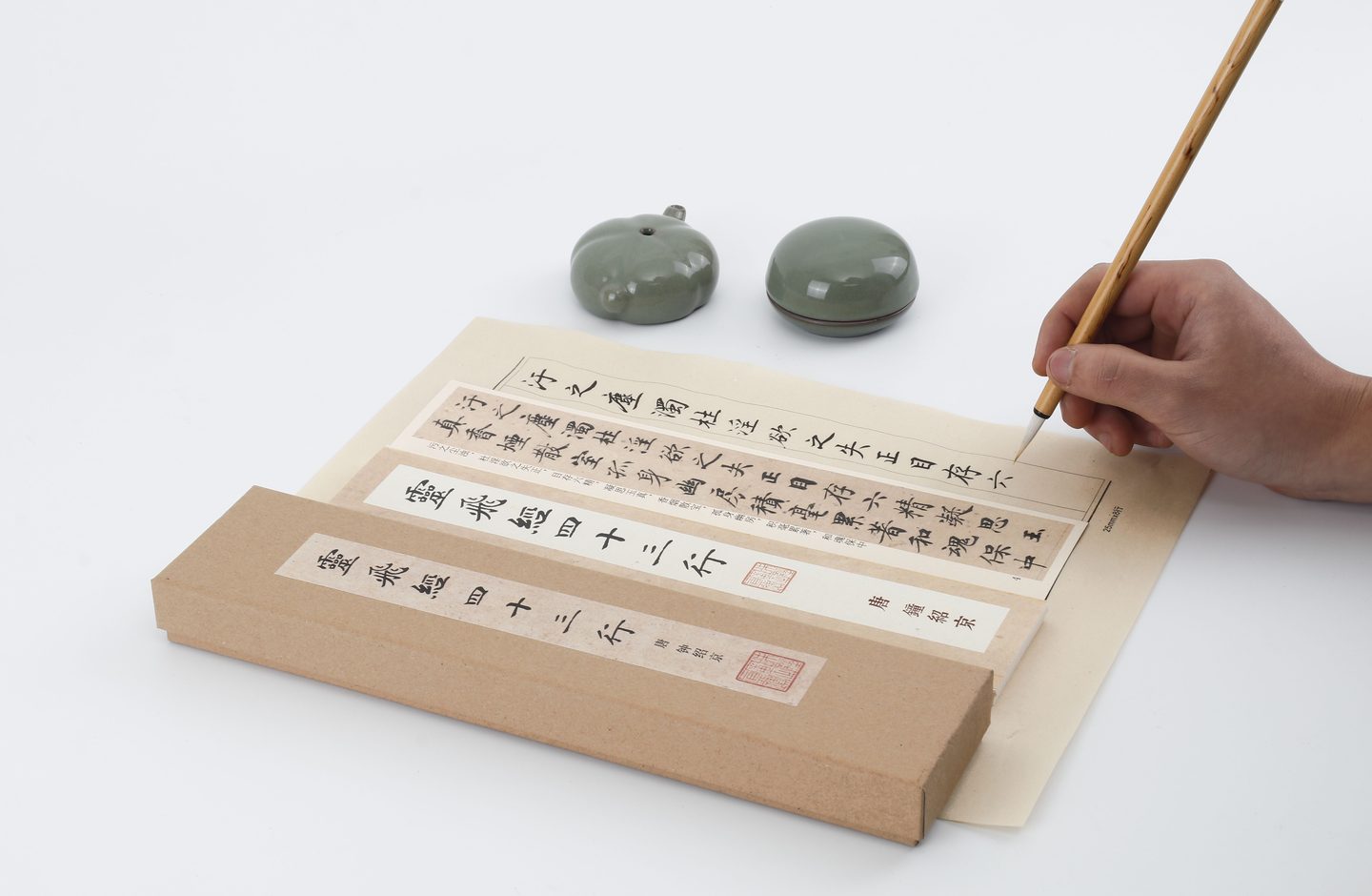 A hand practicing Chinese calligraphy with a brush, copying characters from a printed reference sheet.