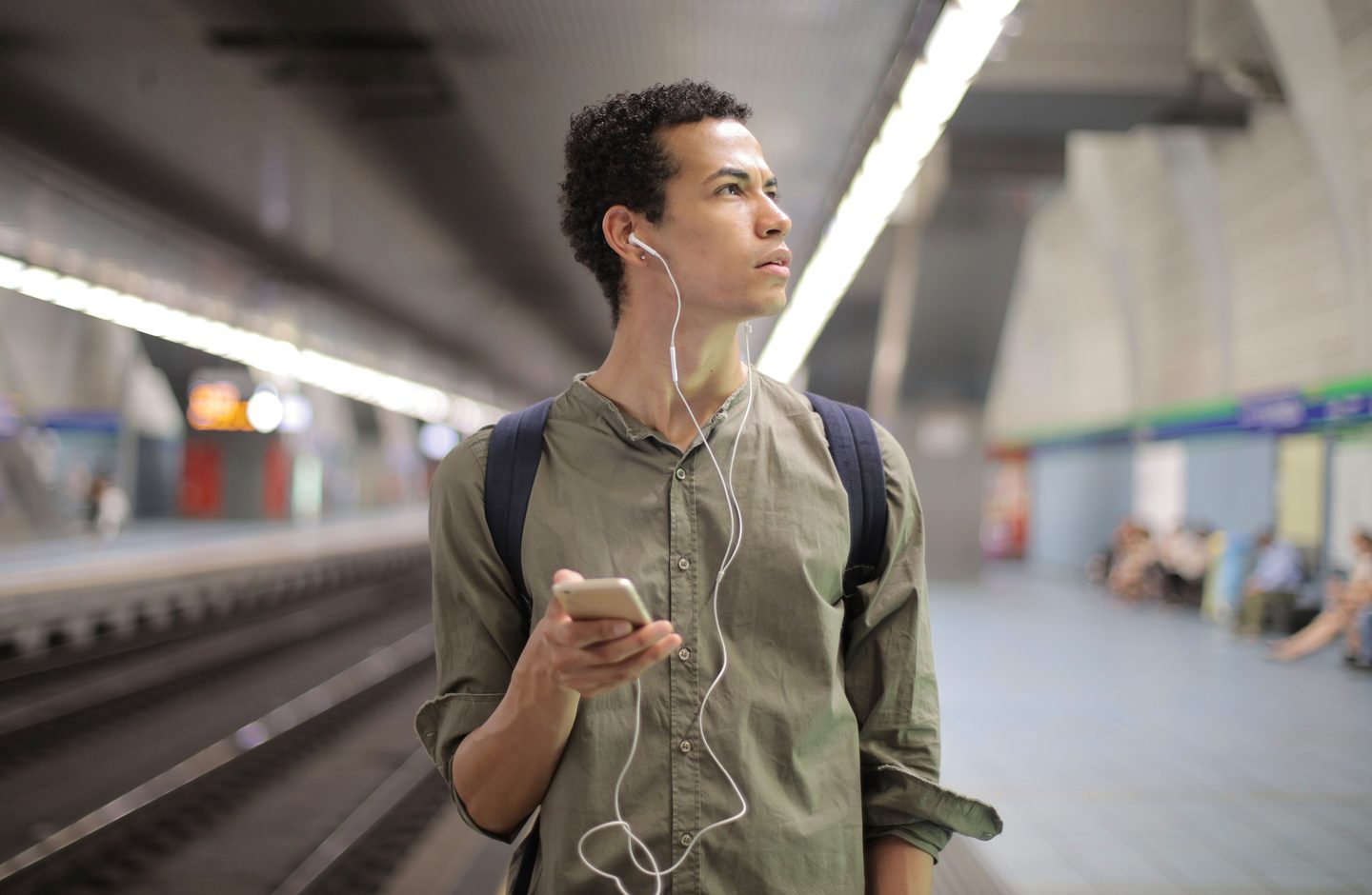 A young man wearing earbuds while waiting at a subway platform, holding his phone.