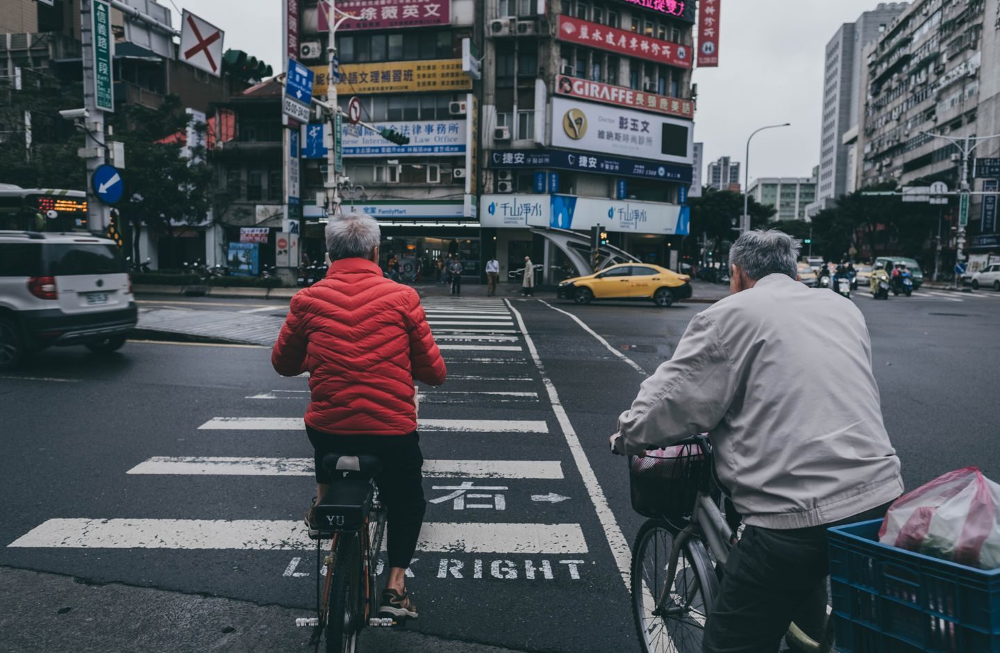 Two elderly men on bicycles crossing a busy Taiwanese street intersection.