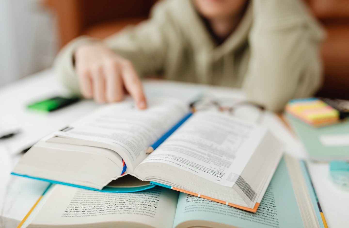A person pointing at a line of text in a thick open reference book on a desk.