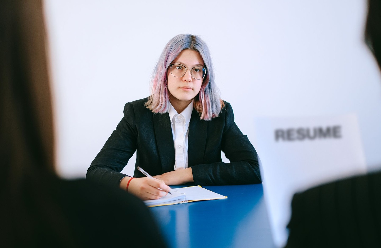 A job applicant in business attire sits across from an interviewer at a desk, with a document labeled Resume visible on the table.