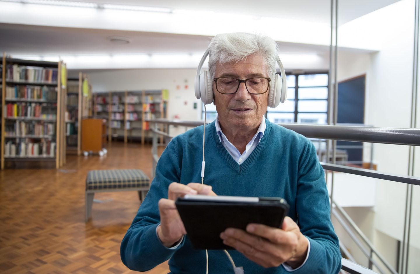 An older man wearing white headphones using a tablet in a public library, with bookshelves behind him.