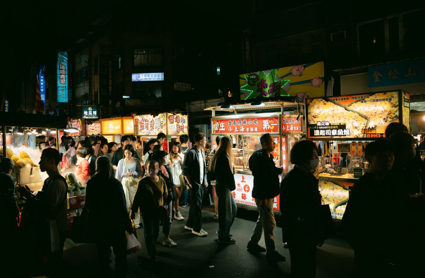 A crowded Taiwanese night market at night, food stalls lit up with signs in Chinese, vendors and customers browsing.