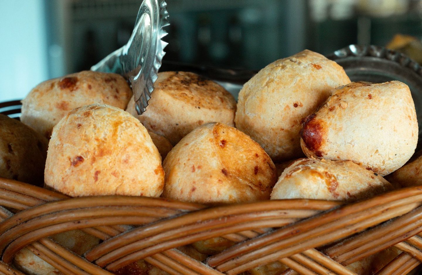 A wicker basket of freshly baked pão de queijo, Brazilian cheese bread, with serving tongs at a bakery in São Paulo.