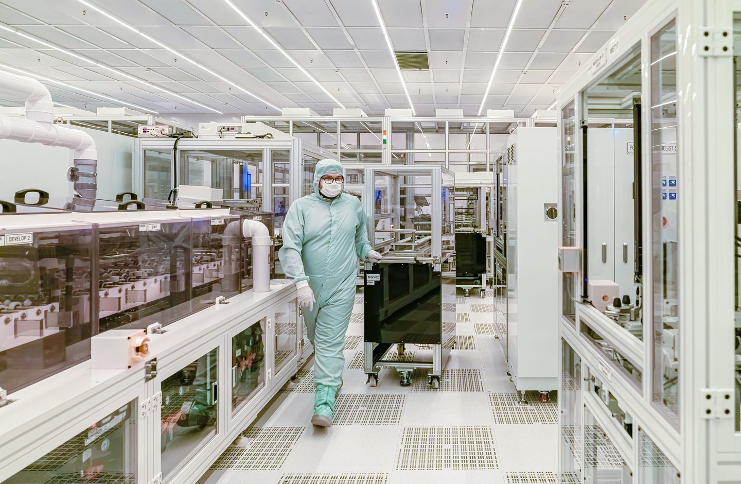 A technician in full cleanroom protective gear walking through a semiconductor fabrication facility lined with processing equipment.