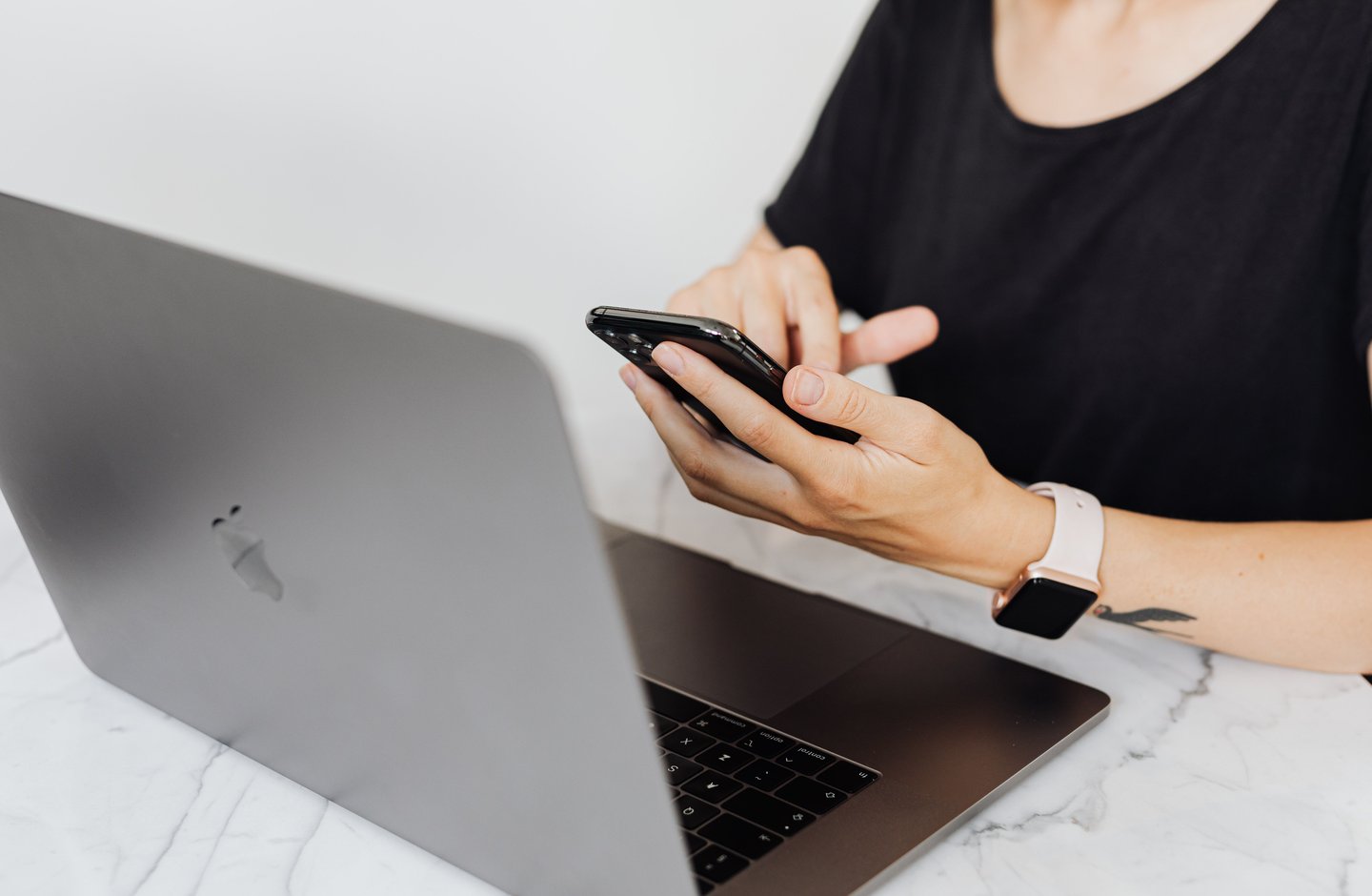 A person holding a smartphone in one hand beside an open laptop on a desk.