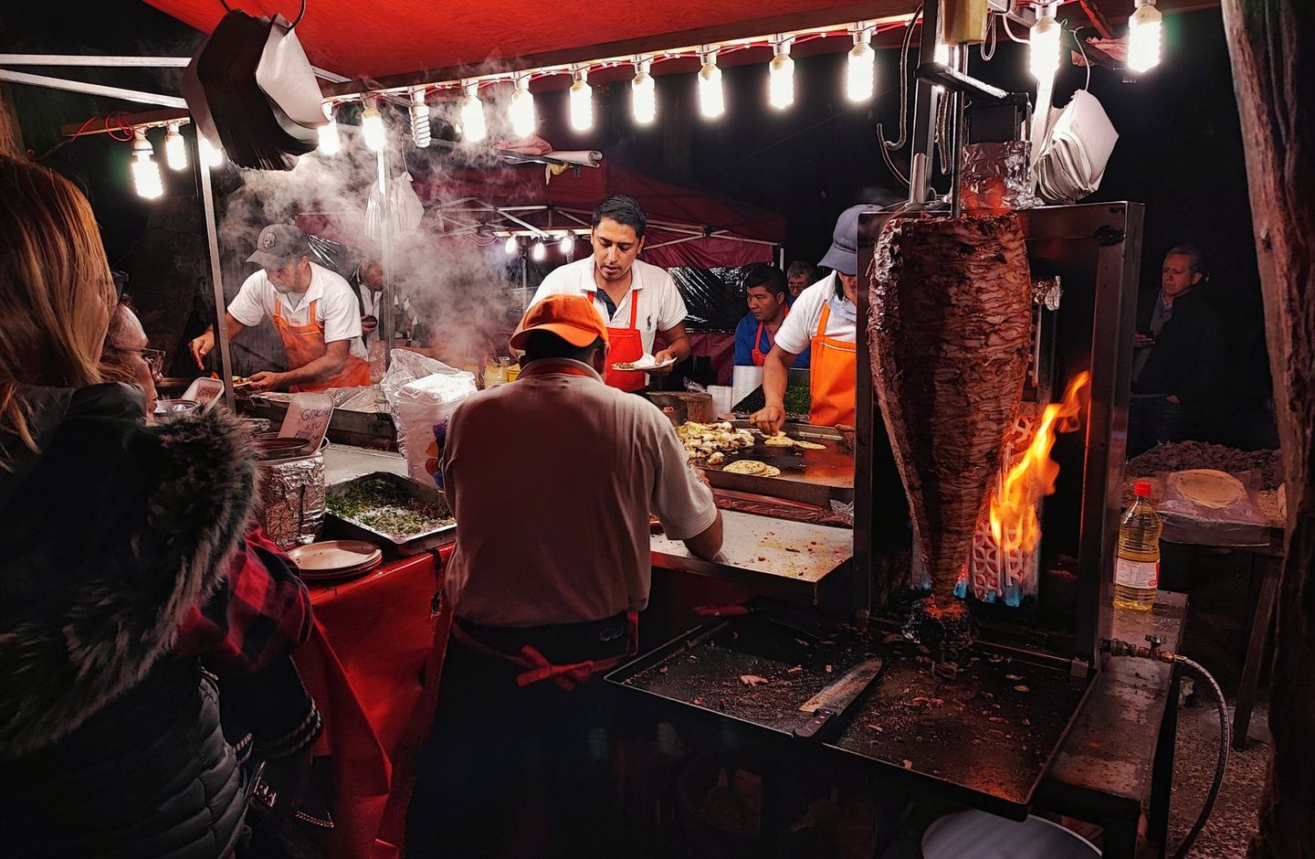 A street taco stand in Mexico City at night, with cooks behind the counter and pork for tacos al pastor turning on a spit.