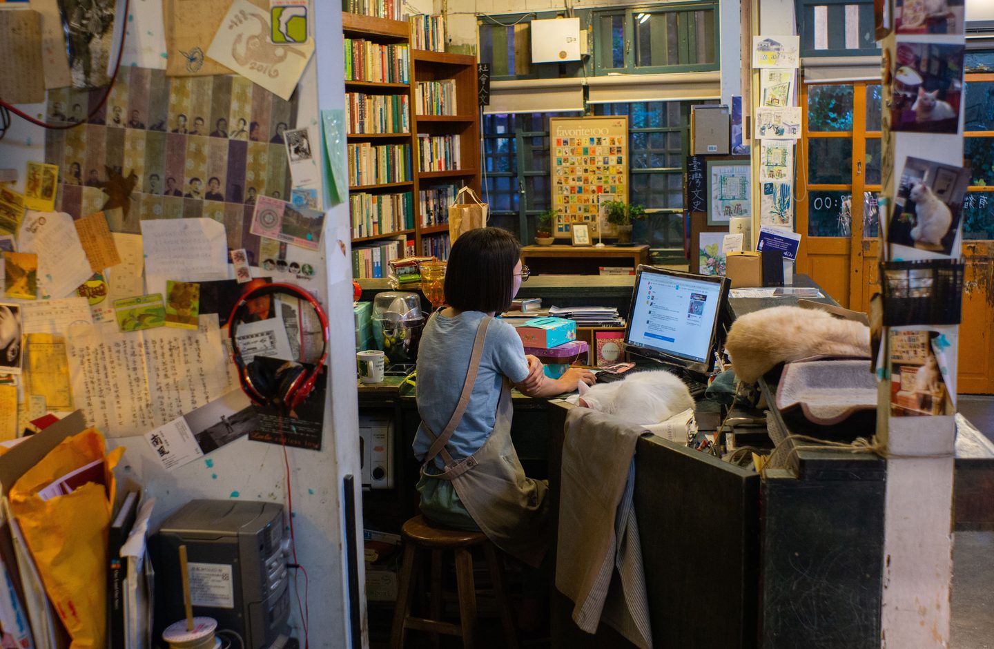 A woman working at a desktop computer behind the counter of an independent Taiwanese bookshop, surrounded by handwritten notes and a hanging sign reading 臺灣文史.