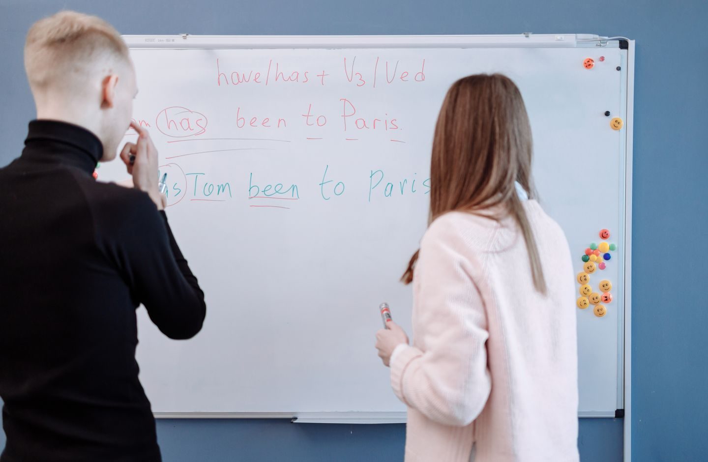 Two people at a whiteboard, one explaining a language lesson to the other.