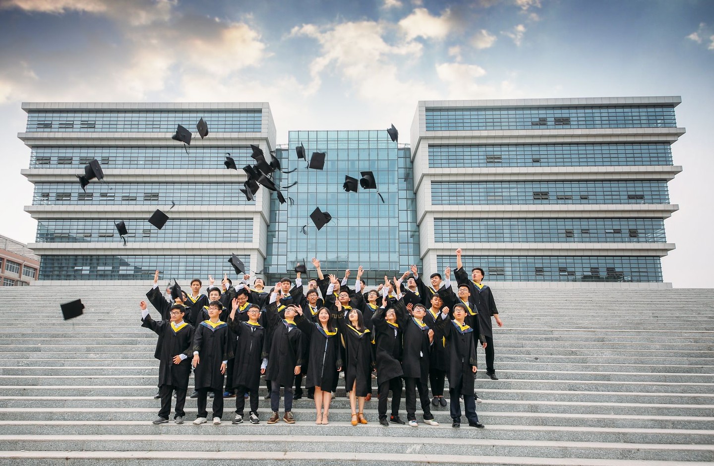 A large group of university graduates in cap and gown throwing their caps in the air in front of a modern glass campus building.