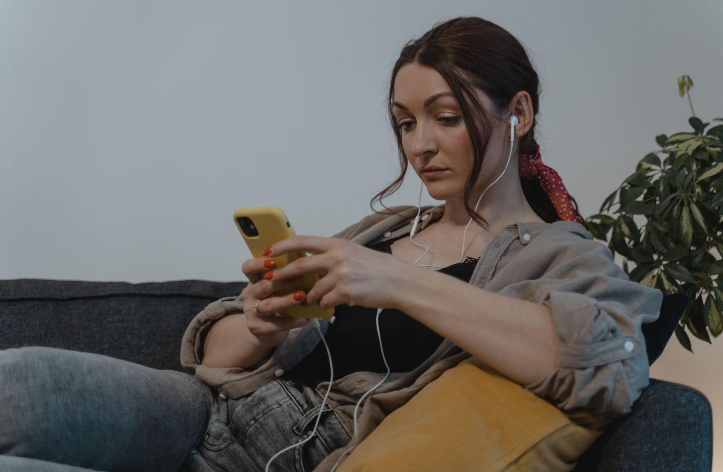 A woman sitting on a couch with wired earphones in, reading something on her phone.