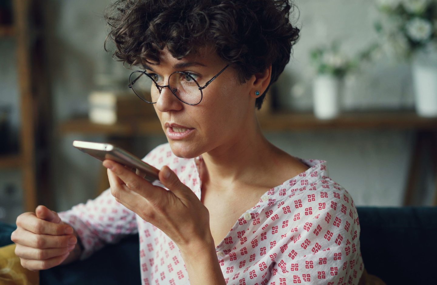 A woman with curly hair and round glasses speaking into her smartphone at home.